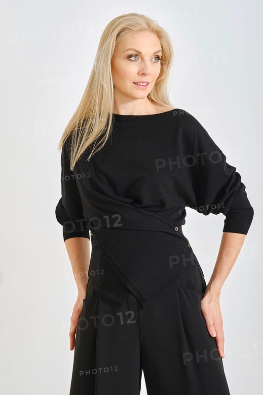 A fashion model stands confidently in a modern black outfit, featuring a stylish wrap design. The studio background highlights her poise and the elegance of the clothing during a photoshoot session