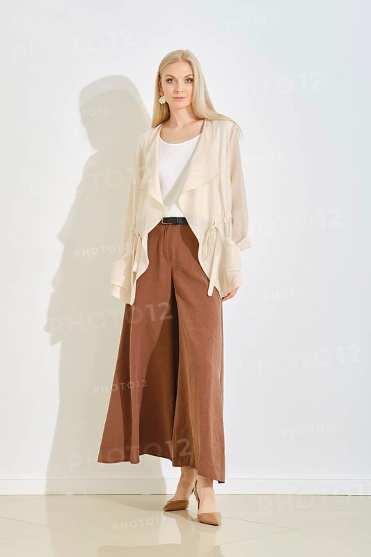 A model displays a fashionable outfit featuring wide, brown pants paired with a lightweight cream blouse. The scene is brightly lit, highlighting the stylish look and clean lines of the clothing