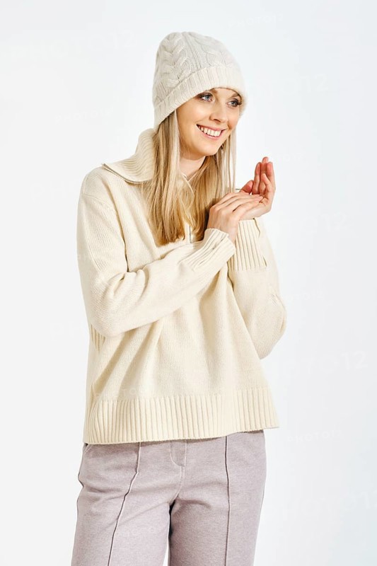 A woman stands with a smile, wearing a sweater and a hat. She clasps her hands together in front of her. The background is white, suggesting indoor lighting. The setting feels cozy and warm