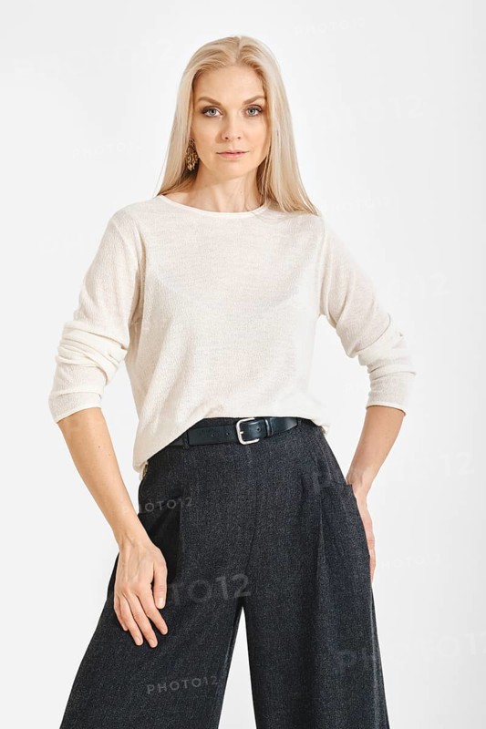 A woman stands with a neutral expression in a studio. She wears a light top and gray wide pants. The background is plain and clear, focusing attention on her outfit and pose