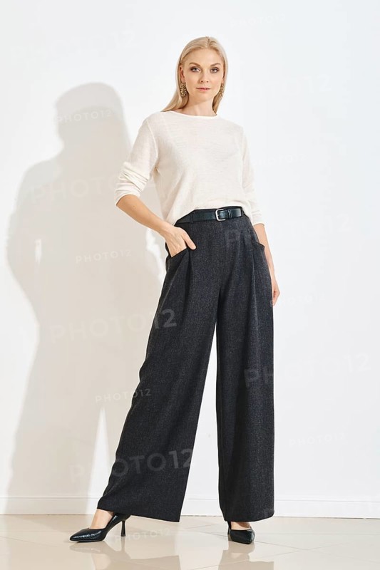A woman in grey pants and a white top poses in a simple indoor space. Her hands are in her pockets. The setting has bright lighting and plain walls, giving focus to her outfit