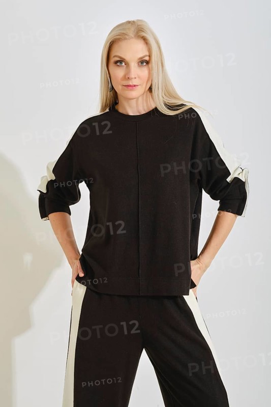 A woman stands with a confident pose wearing a comfortable black tracksuit with white stripes on both sides. The setting features bright lighting, highlighting her relaxed yet fashionable style