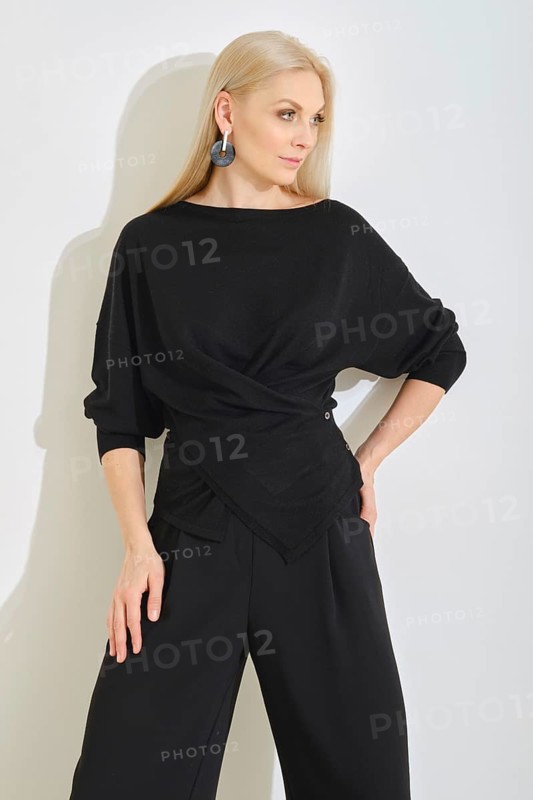 A woman poses gracefully in a modern indoor space, wearing a black wrap top and wide-leg pants. Her elegant style is complemented by statement earrings and soft, flowing hair under natural light