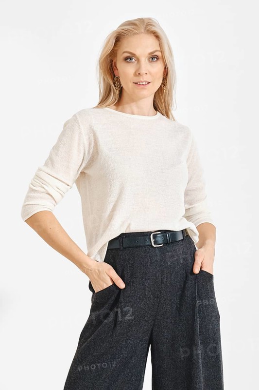 A woman poses with hands in pockets wearing a light shirt and dark pants. She has long blonde hair and stands in a well-lit studio with a simple backdrop. The scene focuses on her confident stance