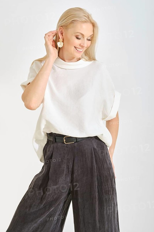 A cheerful woman stands in a bright studio, wearing a simple white top and flowy black pants. She accessorizes with large earrings and showcases a joyful expression while touching her hair