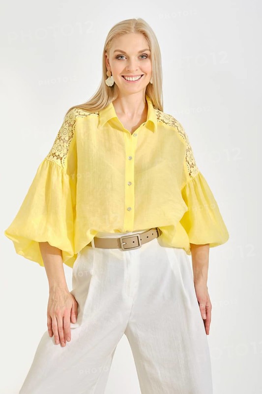 Cheerful woman in a vibrant yellow blouse with lace details and white trousers posing in bright studio