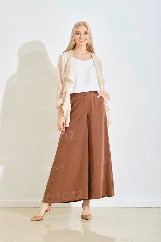 A woman in wide brown pants paired with a light top and a flowing cardigan posing in bright room