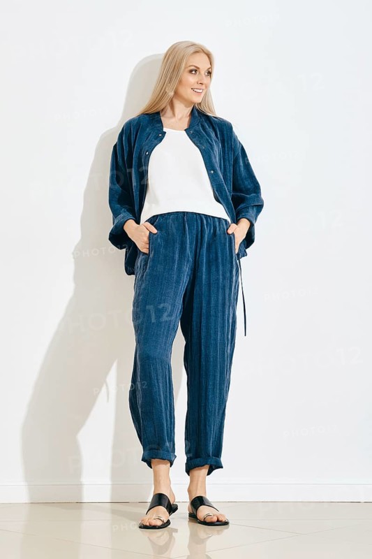 A woman is standing against a white wall in a bright room. She wears a blue pants and jacket with a white top. Her hair is long and straight. The floor has a shiny finish