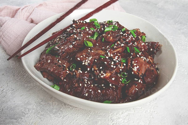 Crispy beef, with green onions and sesame seeds, in a bowl with chopsticks, on a textured surface, on a light background, no people
