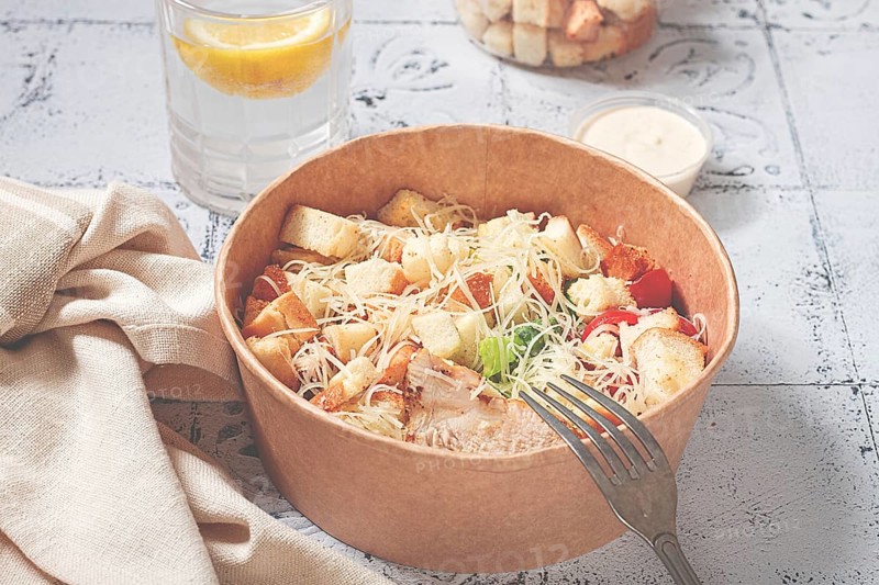 Caesar salad in a paper container, take it on the way, with sauce and croutons, on the table, snack, natural light, no people