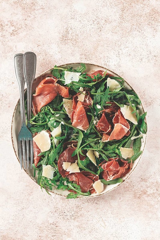 Fresh arugula salad with prosciutto and grated parmesan cheese, on a plate, homemade, no people