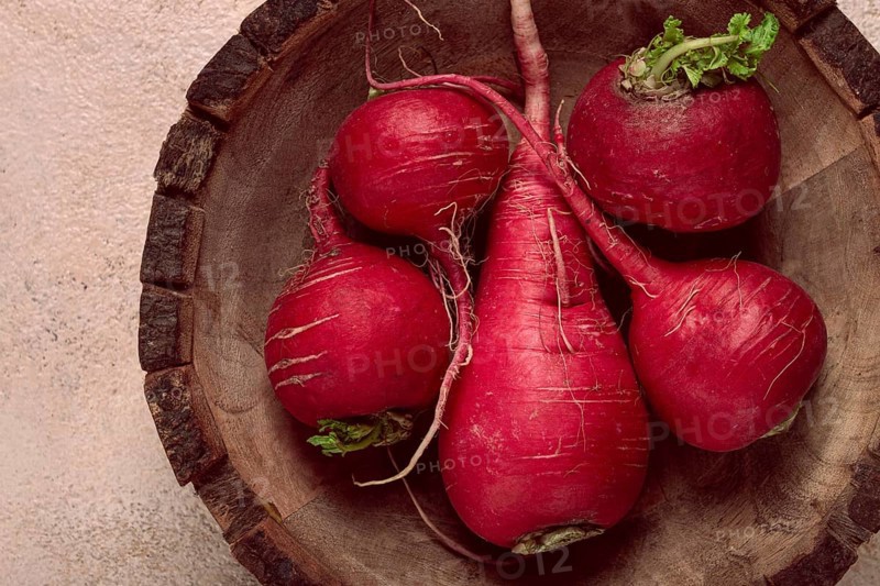 A collection of vibrant red radishes sits in a round wooden bowl, showcasing their natural texture and earthy tones against a softly colored background