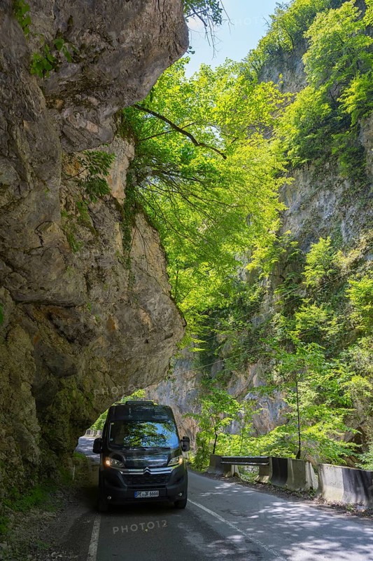 A black van drives along a shady mountain road lined with rocks and trees, camper, motorhome in a gorge on the Lajanuri river near Achara, Racha-Lechkhumi region and Lower Svaneti, Georgia