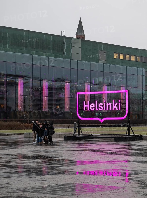 Tourists with selfie sticks take pictures of themselves in front of the glowing pink Helsinki city logo, square in front of Oodi Central Library and Cultural Center, Helsinki, Finland