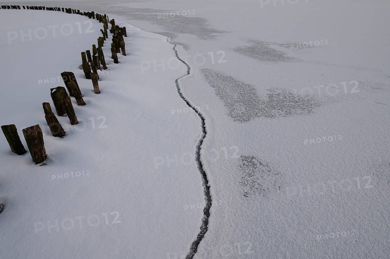 Frozen lake surface of Dümmer See with wooden posts and distinctive fault line in ice, frozen landscape with cracks and barriers on ice under grey sky, Lembruch, Diepholz district, Lower Saxony, Germany