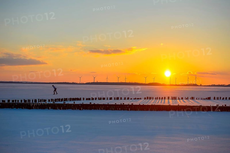 Ice skaters in front of sunset over icy landscape with wind turbines and intense colors on the frozen Dümmer Lake, Lembruch, Diepholz district, Lower Saxony, Germany