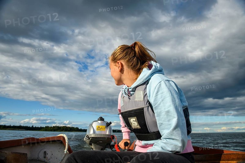 Young woman in a boat with outboard on the quiet lake Vänern, looking into the distance, the sky is cloudy, Vänern, Värmland, Sweden