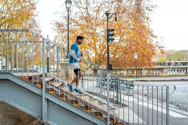 Energetic black man maintaining fitness by running down concrete stairs in an urban setting with autumn trees and a bridge in paris, demonstrating an active, healthy city lifestyle