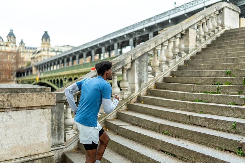 Young black man engaging in a fitness workout, actively running up historical stone stairs in paris with a bridge and buildings visible in the urban background
