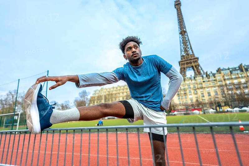 Young black man stretching legs on a paris running track before a workout, eiffel tower and haussmann buildings in the background, focused on urban fitness and dedication
