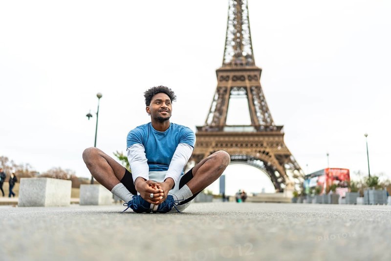 Young black man doing a butterfly stretch on paris pavement, warming up for urban run with the eiffel tower in the background, focused and smiling, active lifestyle