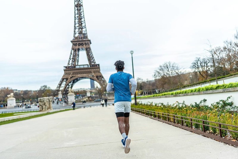 Young man in athletic wear jogging through a park pathway with the iconic eiffel tower prominently featured in the background on a cloudy day, focusing on fitness and tourism
