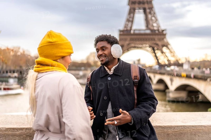 Diverse couple enjoying a romantic winter getaway in paris on a bridge by the eiffel tower, chatting and smiling as they sightsee along the seine during a city break