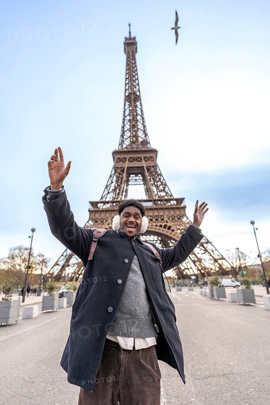 Young man with winter earmuffs raising arms in excitement and smiling widely while visiting the eiffel tower in paris, celebrating travel and tourism on a fun trip