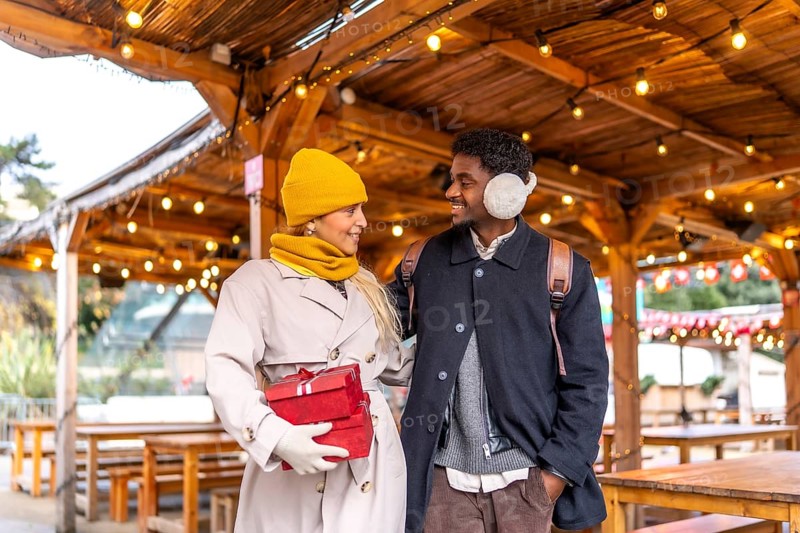 Diverse couple walking together and exchanging smiles in a festive outdoor christmas market in paris, carrying gifts and celebrating the holiday season