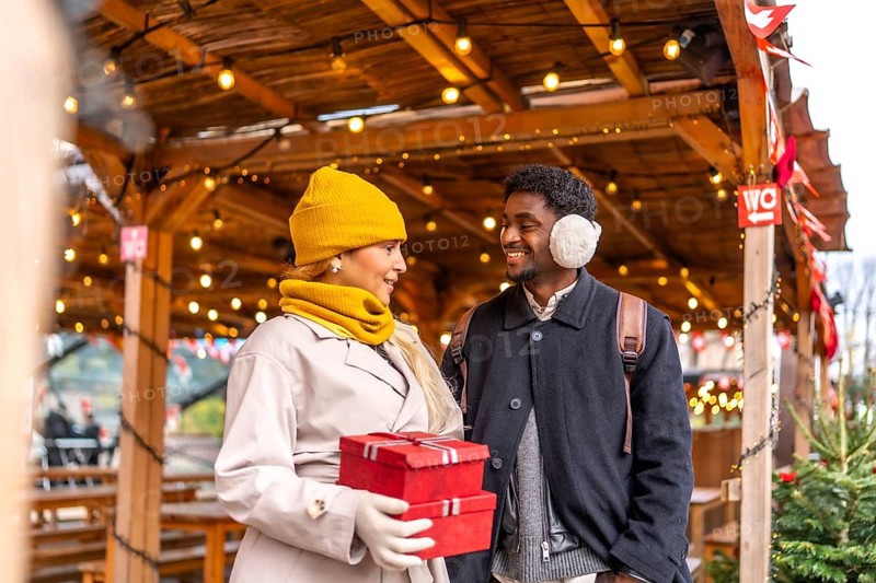 Young couple smiling at each other, walking and holding presents in red boxes at a festive outdoor christmas market in paris, enjoying a winter holiday together