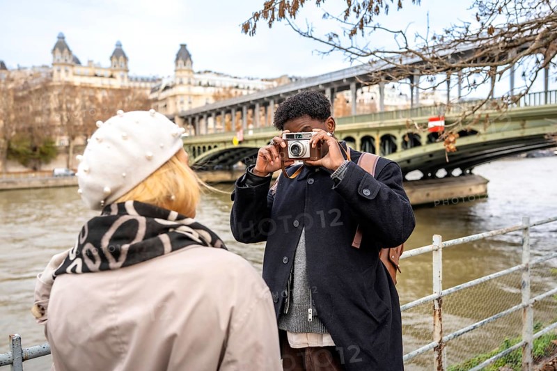 Young man photographing his partner with a vintage camera while exploring the city of paris with the bir hakeim bridge and iconic architecture in the background on an overcast day