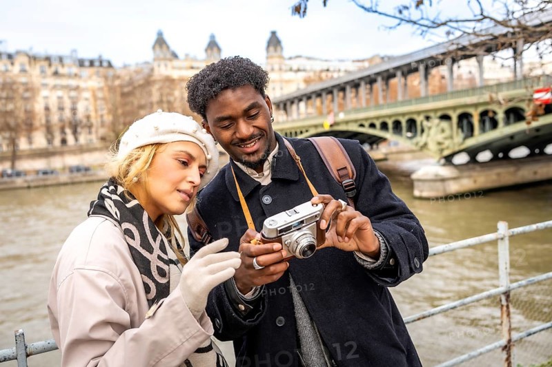 Young multiracial travelers enjoying a romantic city trip to paris, standing by the seine river and sharing a moment while looking at a vintage camera
