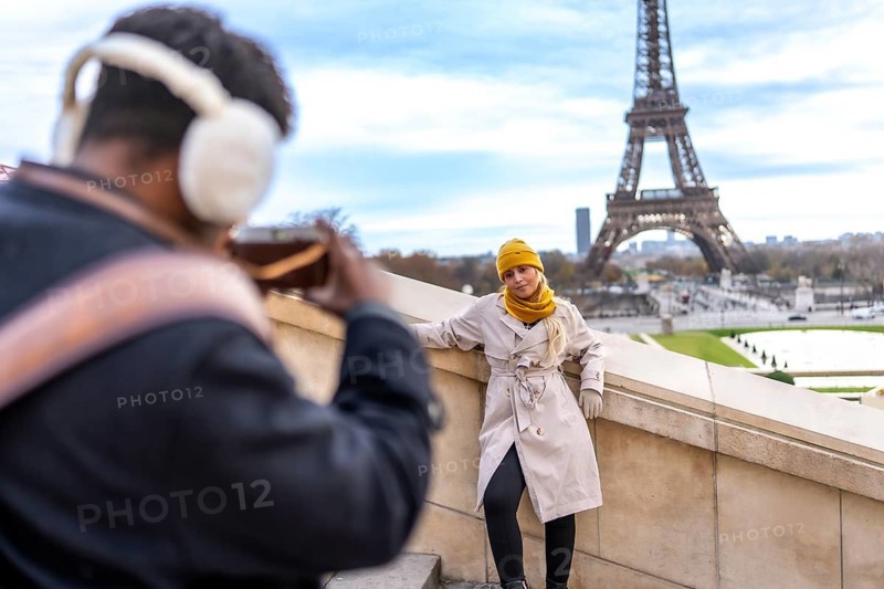 Couple traveling in paris. France. With a man taking a smartphone picture of his blonde girlfriend posing on a stone wall overlooking the iconic eiffel tower on a cloudy day. Creating travel memories