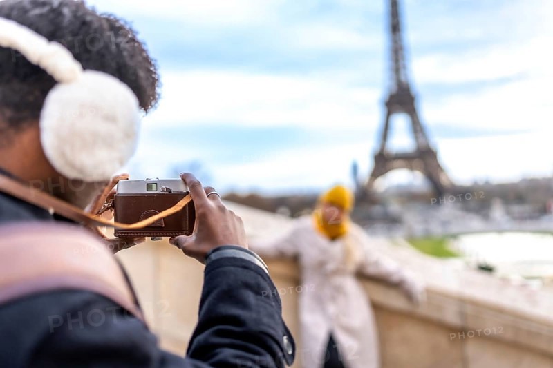 Young man wearing earmuffs and using a vintage camera to photograph his female partner posing with the iconic eiffel tower in the background, enjoying a romantic trip to a european city