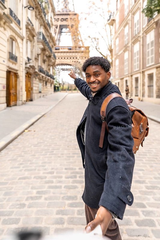 Happy black man leading someone by the hand down a cobblestone paris street, smiling and pointing toward the eiffel tower while enjoying a carefree travel moment