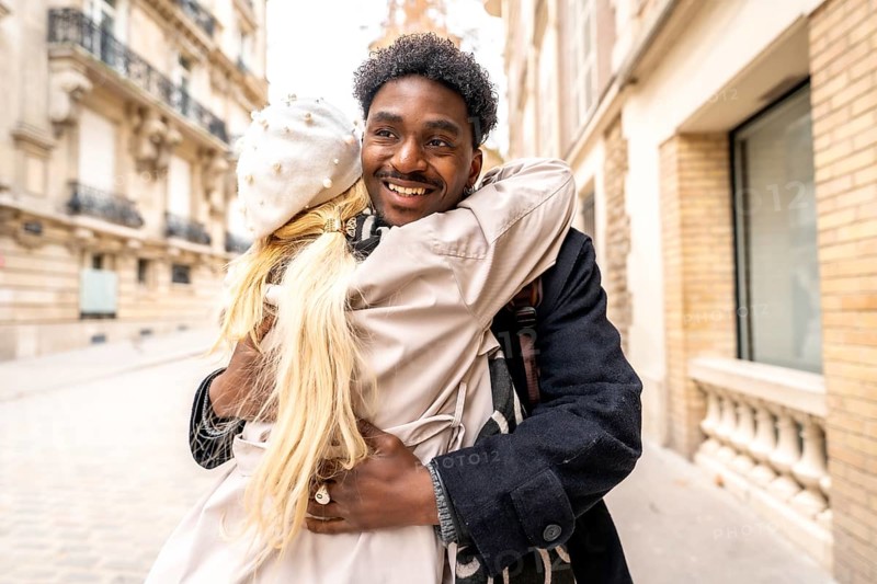 Loving diverse couple embracing and smiling on a charming parisian street, sharing a warm, romantic moment during an autumn stroll amid classic architecture and cozy fashion