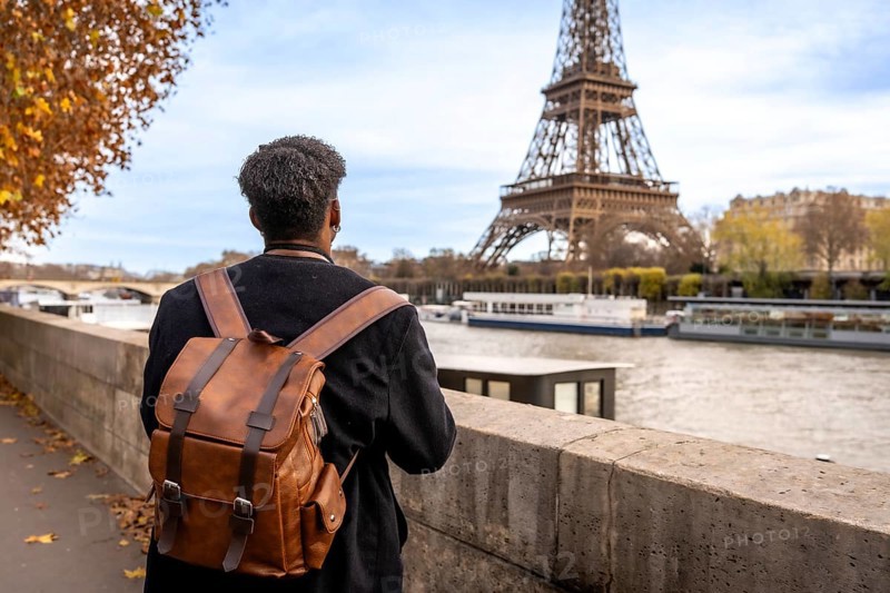 Person with travel backpack standing by the seine, gazing at the eiffel tower amid autumn foliage on a solo city trip in paris, enjoying urban riverside views and light breeze