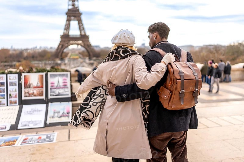 Young diverse couple embracing on a paris street, back view toward the eiffel tower and trocadero, enjoying a romantic sightseeing stroll amid artists' stalls and autumn coats