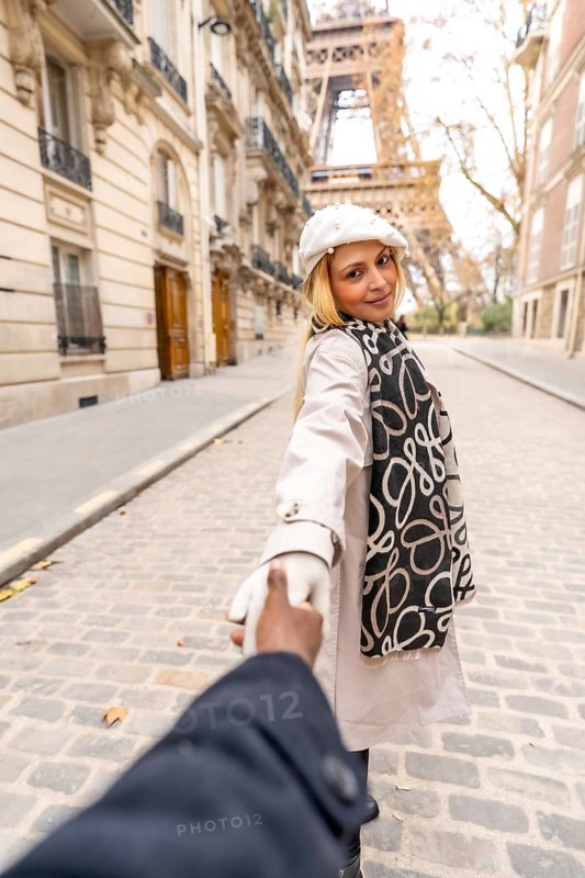 Woman holding her partner's hand, smiling and looking back while walking on a cobblestone paris street with haussmann buildings and the eiffel tower in the background, romantic travel vibe