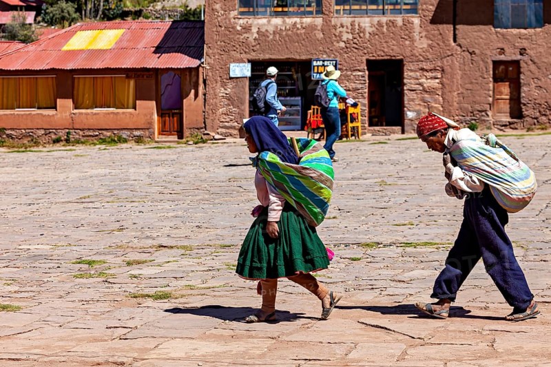Two woman in traditional clothing carry bundles on their backs through a village in Bolivia in daylight, farmers walk across a square on the island of Taquile in Lake Titicaca in Peru