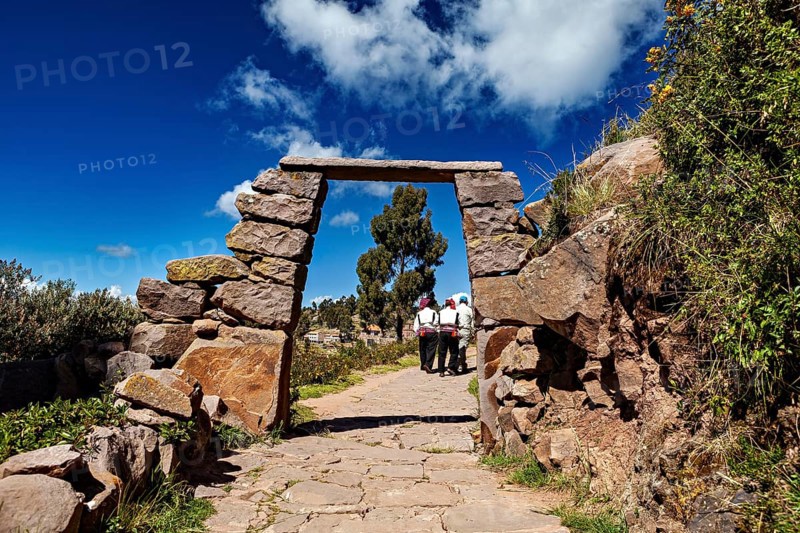 People wearing traditional clothes walk under a rectangular stone arch on a rural path, stone gates on Taquile Island in Lake Titicaca in Peru
