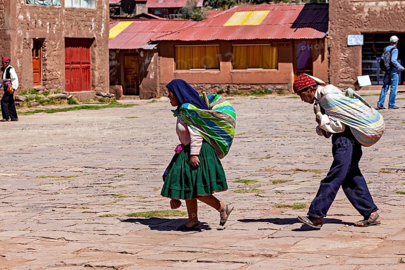 Two woman in colorful clothes carry loads on their backs and walk through a sunny village in Bolivia, farmers walk across a square on the island of Taquile in Lake Titicaca in Peru