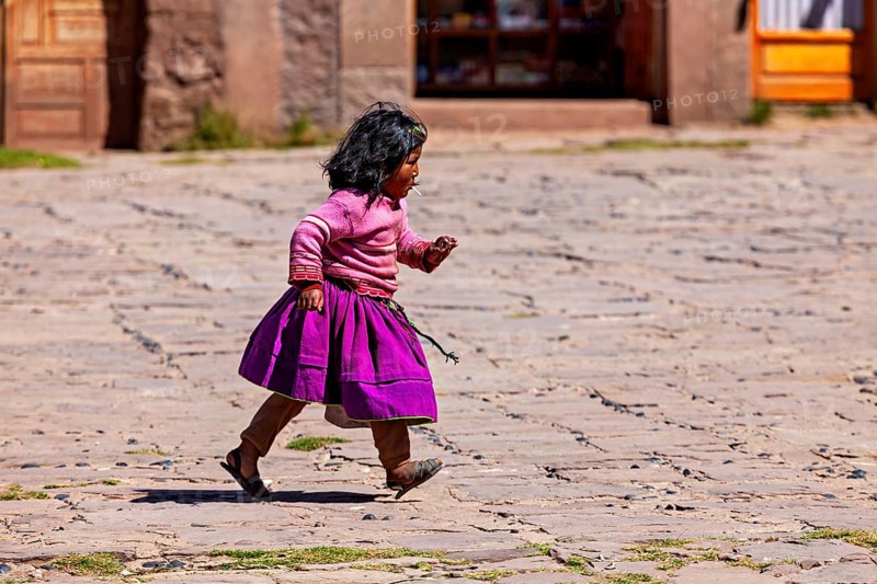 Little girl in purple skirt and pink sweater walks across a paved square, children from Taquile Island in Lake Titicaca in Peru