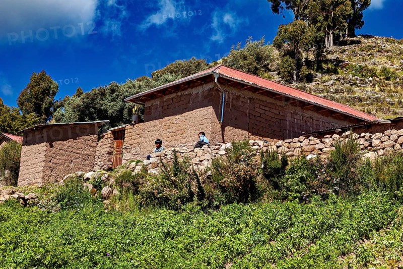 Two people in front of rustic farm houses surrounded by green fields under a blue sky, the countryside and farmhouses on Taquile Island in Lake Titicaca in Peru