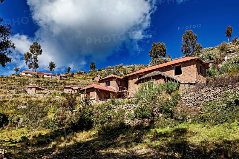 Houses cluster on a hillside under a dramatically cloudy sky framed by trees and vegetation, The countryside and farmhouses on the island of Taquile in Lake Titicaca in Peru