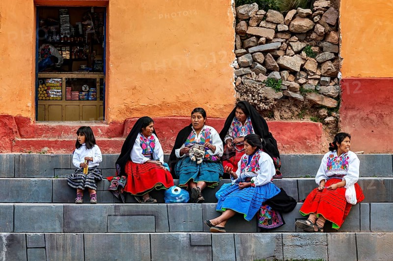 Women and girls in colorful clothes sit on stairs in front of a complex of buildings, The woman on Taquile Island in Lake Titicaca in Peru