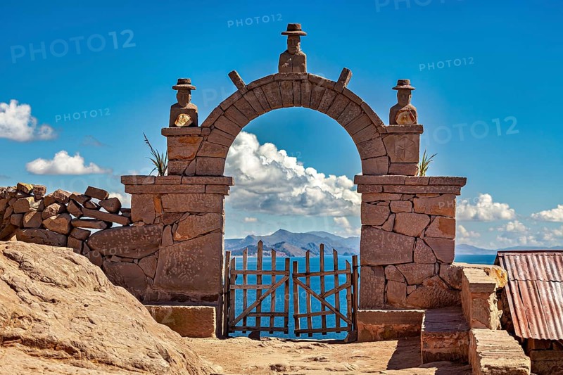 A traditional stone arch with wooden slats stands in front of a distant mountain landscape under clear blue skies, stone gates on the island of Taquile in Lake Titicaca in Peru