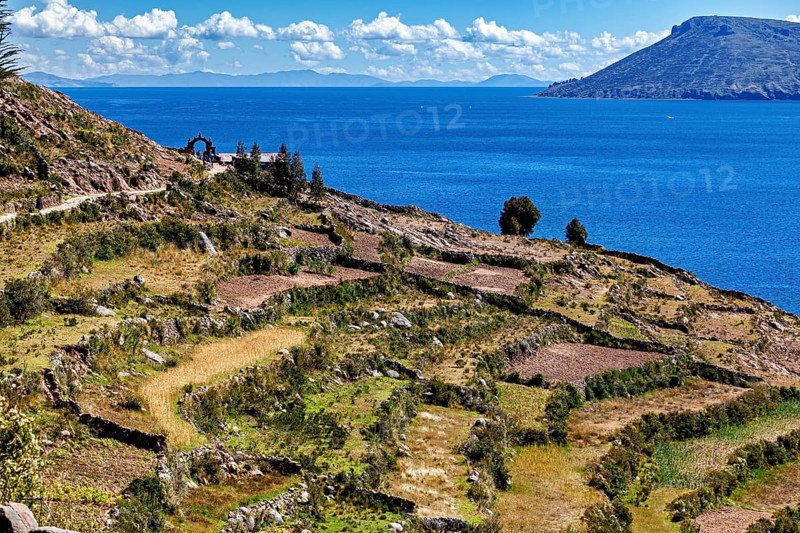 Terraced fields stretch down to a clear blue lake, framed by stone arches and hills, the countryside and farmhouses on the island of Taquile in Lake Titicaca in Peru