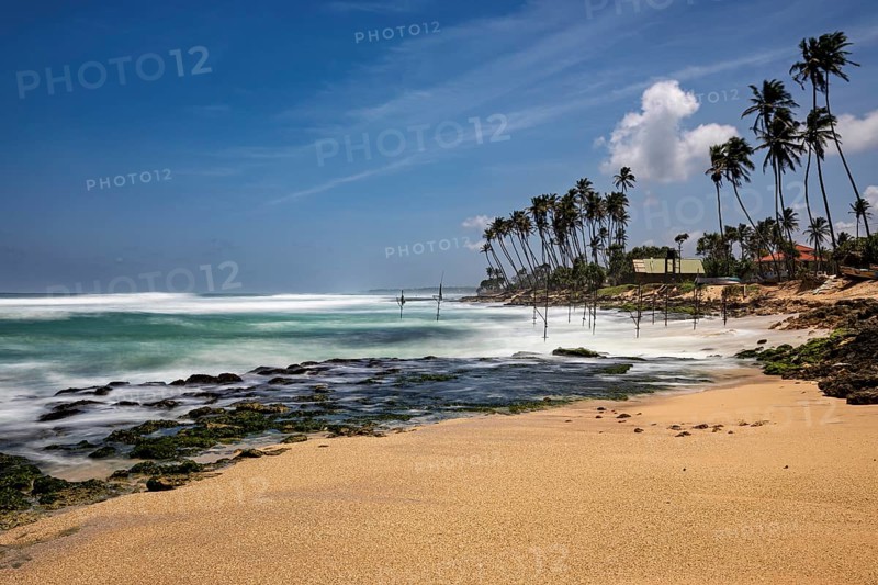 A sun-drenched beach with palm trees and waves hitting the rocks, Koggala beach with stilt fishermen in Sri Lanka