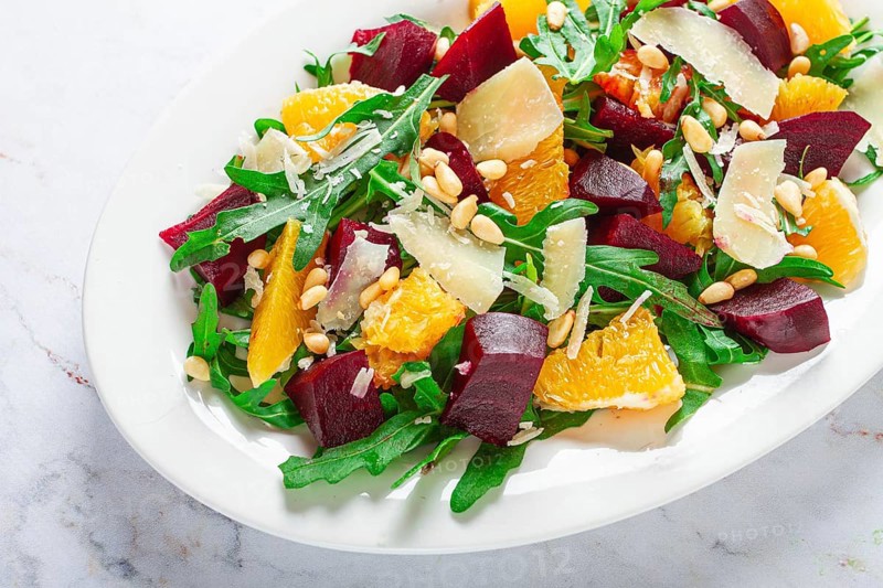 A salad made with chunks of beet, orange slices, and arugula. It is topped with cheese shavings and pine nuts, placed on a white plate with a striped napkin beside it